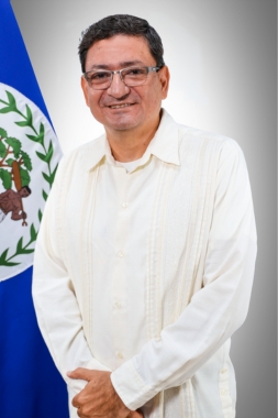 Dr. Louis Zabaneh, in a cream guayabera, beside the flag of Belize